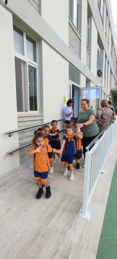 Uczniowie wraz z nauczycielami opuszczają budynek szkoły. Uczniowie ubrani są w szkolne mundurki w kolorze pomarańczowym.