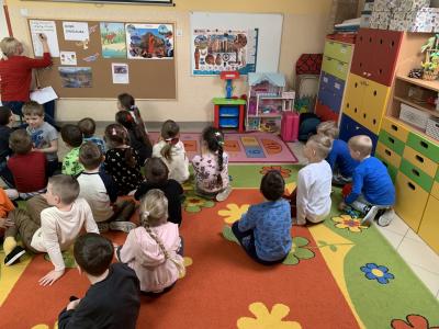 Dzieci siedzą na dywanie w sali przedszkolnej przed tablicą korkową, na której widnieje napis ,,Dzień dinozaura, przypięte są różne sylwet dinozaurów.