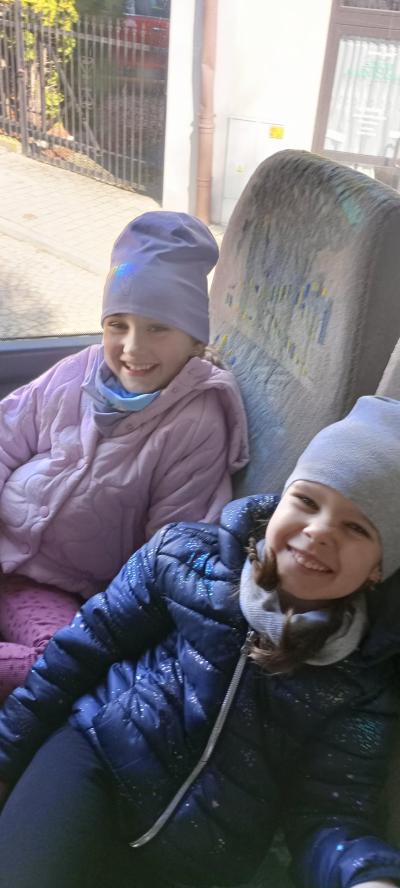 Dwie dzieczynki z szerokim uśmiechem na twarzy patrzą w stronę fotografa, siedząc na fotelach w autobusie