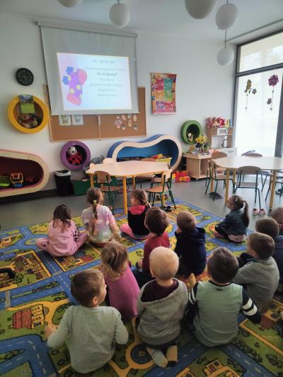 Dzieci siedzą na dywanie przed ekranem. Oglądają bajkę edukacyjną. Na ekranie fioletowa skarpetka.