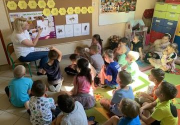 Dzieci siedzą przed nauczycielem i tablicą tematyczną który pokazuje ilustracje z  cyklem rozwojowym pszczół.