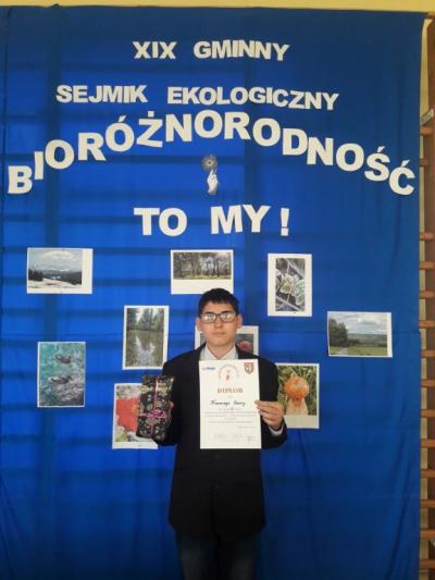 Uczeń klasy 8b Ksawery Szary z dyplomem i nagrodą za zajęcie III miejsca w konkursie fotograficznym. W tyle hasło tegorocznego sejmiku i zdjęcia ukazujące bioróżnorodność przyrody.