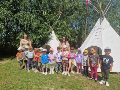 Grupa dzieci wraz z dwoma paniami pozuje do zdjęcia. Za nimi drzewa i 2 indiańskie namioty tipi..JPG