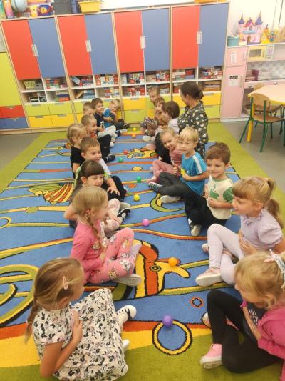 Dzieci siedzą na dywanie naprzeciwko siebie w dwóch rzędach. Pomiedzy każdą parą dzieci leży kolorowa piłeczką..jpg