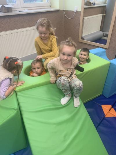 3 dziewczynki siedzą na zielonych dużych geometrycznych pufach, a 2  chłopcy schowali się do otworów w nich ..jpg