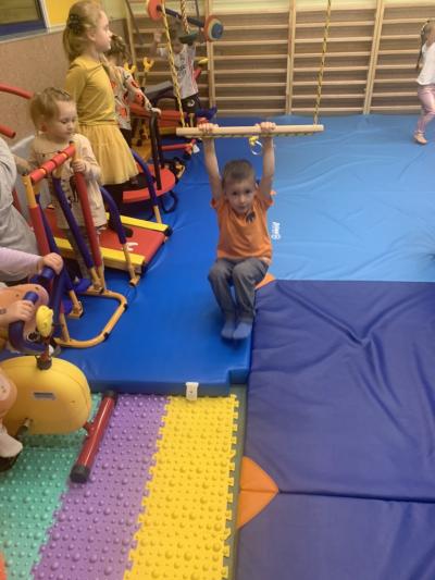 Chłopiec kołysze się trzymając drążek na linach a reszta dzieci ćwiczy na mini przyrządach w sali ćwiczeń..jpg
