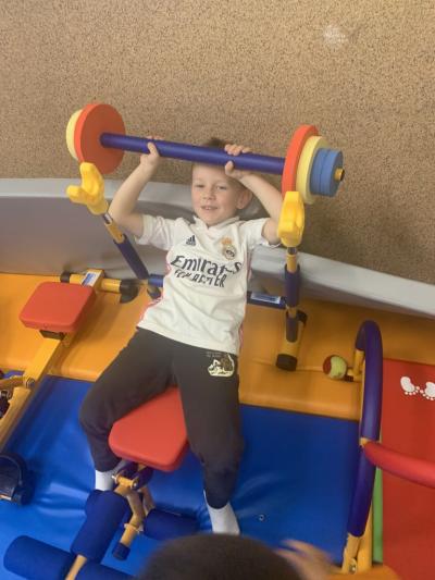 Chłopiec leży na mini przyrządzie a w dłoniach trzyma mini sztangę w sali ćwiczeń..jpg