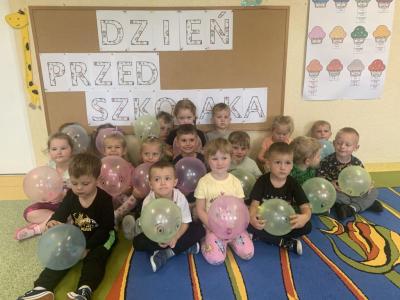 Dzieci siedzą na dywanie przed tablicą z napisem ,,Dzień przedszkolaka i trzymają w dłoniach nadmuchane balony..jpg