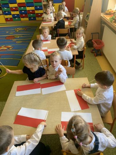 Dzieci siedzą przy stolikach i wykonują flagi z białego i czerwonego papieru oraz długiej wykałaczki..jpg