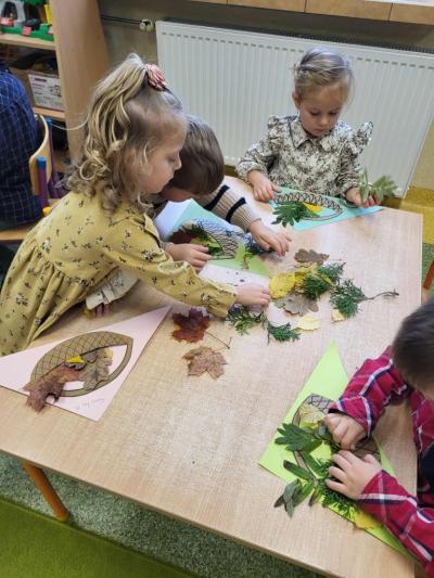 Dzieci siedzą przy stoliku i wkładają suche liście do tekturowego koszyczka naklejonego na kartkę..JPG