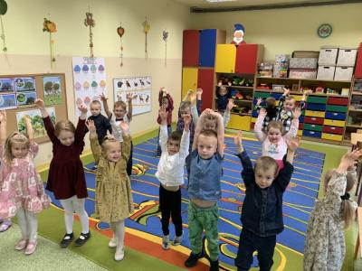Dzieci stoją na dywanie w sali przedszkolnej z podniesionymi do góry rękami..jpg
