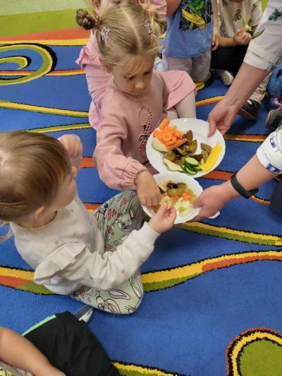 Nauczyciel częstuje siedzące na dywanie dzieci pokrojonymi na talerzu owocami..JPG