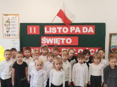 Dzieci z grupy Sówki stoją w postawie na baczność do odśpiewania hymnu. W tle tablica z  dekoracją..jpg