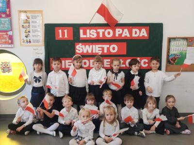 Zdjęcie grupowe dzieci z grupy Sówki. Dzieci trzymają flagi. W tle tablica z dekoracją..jpg