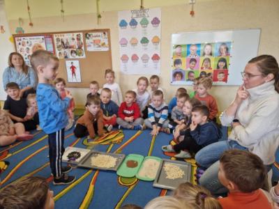 Chłopiec stoi przed zgromadzonymi na dywanie tackami z różnymi rodzajami ziaren. Reszta dzieci z grupy wraz z nauczycielem siedzą w kole..JPG