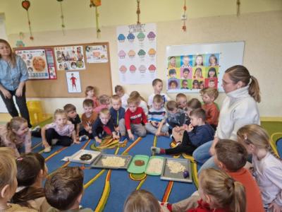 Dzieci siedzą wraz z nauczycielem w kole na dywanie. W środku koła leżą zgromadzone tacki z różnymi rodzajami ziaren..JPG