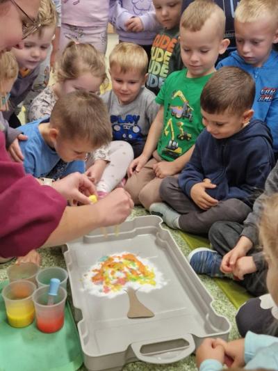 Dzieci zgromadzone przed nauczycielem przyglądają się z zaciekawieniem eksperymentom z pipetą i kolorowymi farbami które barwią wacikową koronę drzewa..JPG