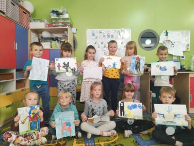 Wspólne zdjęcie dzieci z grupy Skrzaty wraz z pracami plastycznymi. W tle tablica z dekoracją z okazji Dnia Postaci z Bajek.