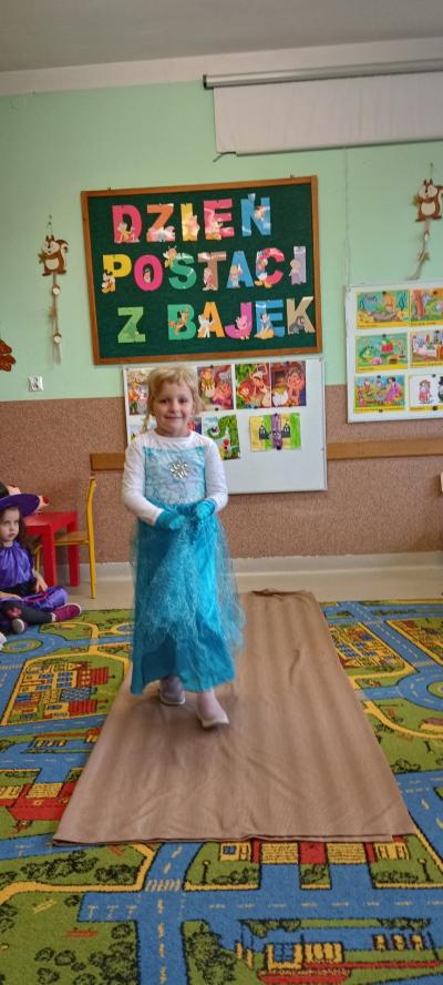 Uśmiechnięta dziewczynka w przebraniu księżniczki Elsy idzie po dywanie. Za plecami dziewczynki widnieje napis Dzień Postaci z Bajek oraz obrazki z bajkowymi postaciami