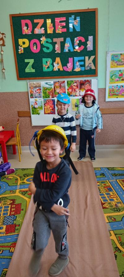 Trzech chłopców z uśmiechniętymi minami i kaskami na głowach  idą gęsiego po dywanie.  Za ich plecami widnieje napis Dzień Postaci z Bajek oraz obrazki z bajkowymi postaciami.