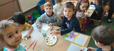 Grupa wesołych dzieci siedzących przy stoliku maluje farbami pracę plastyczną. Trzymają w rękach pędzle, a na środku stołu położone są palety z farbami i kubeczek z wodą.
