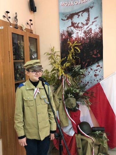 zaŚpiewaj i Ty Niepodległej - chłopiec ubrany w mundur harcerski, stoi przed mikrofonem. Z tylu dekoracja patriotyczna- polska flaga, zwisająca biało-czerwona wstążka, biało- czerwone wrzosy