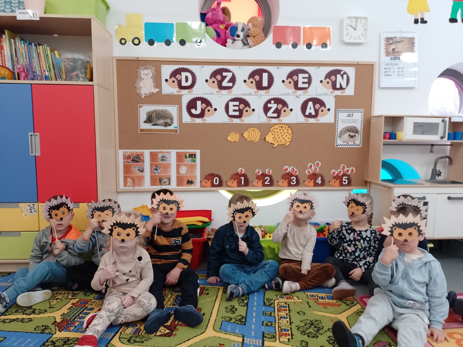 Grupa dzieci siedzi w sali przedszkolnej na kolorowym dywanie. Dzieci trzymają w ręku maski jeża i przykładają je do swoich oczu patrząc przez nie w stronę aparatu. Z tyłu w tle widać tablicę  z napisem dzień jeża oraz obrazki przedstawiające jeże. Nad ta