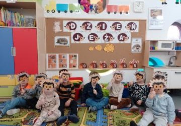 Grupa dzieci siedzi w sali przedszkolnej na kolorowym dywanie. Dzieci trzymają w ręku maski jeża i przykładają je do swoich oczu patrząc przez nie w stronę aparatu. Z tyłu w tle widać tablicę  z napisem dzień jeża oraz obrazki przedstawiające jeże. Nad ta