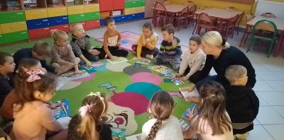 Dzieci siedzą w kole na dywanie i wystukują rytm na kartce papieru do piosenki „Kolorowe kredki”