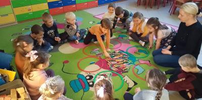 Dzieci siedzą na dywanie w kole razem z nauczycielem i podnoszą do góry taki kolor kredki jaki kolor kartoniku nauczyciel podniósł do góry.