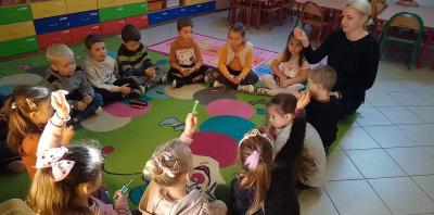Dzieci siedzą na dywanie w kole razem z nauczycielem i podnoszą do góry taki kolor kredki jaki kolor kartoniku nauczyciel podniósł do góry.