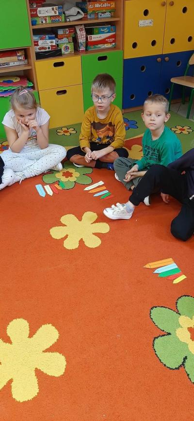 Dzieci z grupy Skrzatów i Pszczółek siedzą na dywanie i układają szablony kolorowych kredek od najmniejszej do największej i odwrotnie.