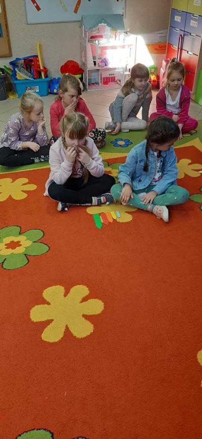 Dzieci z grupy Skrzaty i Pszczółki siedzą na dywanie i układają kredki do słów refrenu piosenki „Kolorowy deszcz”.