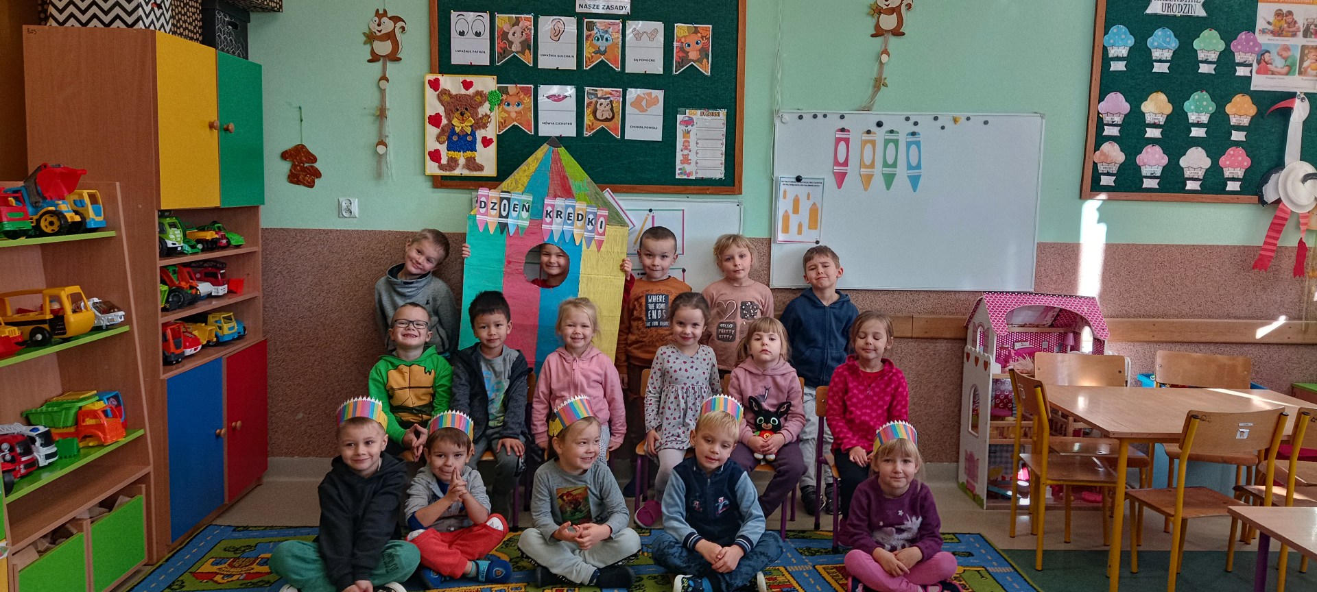 Dzieci z grupy „Leśnych Duszków” stoją w trzech rzędach przy tablicy. Z tyłu za nimi widać tablice z różnymi obrazkami.
