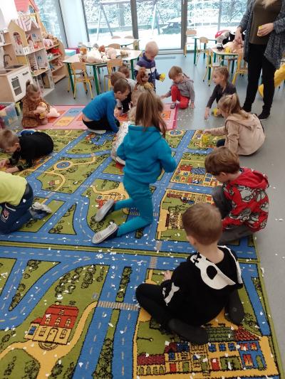 Dzieci kucają na dywanie i zbierają okruszki papierków, które rozsypały. W tle widać stoliki i krzesełka oraz okna.