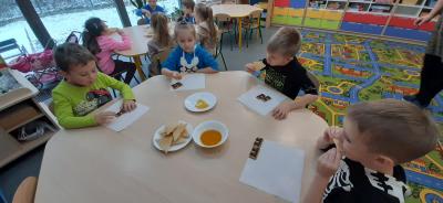 Dzieci z grupy Skrzaty siedzą przy stolikach i degustują miód oraz misiowe ciasteczka