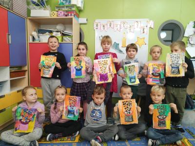 Wspólne zdjęcie dzieci z grupy Skrzaty wraz z pracami plastycznymi. W tle tablica z dekoracją z okazji Andrzejek.