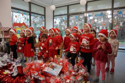 Przedszkolaki ubrane w czerwone stroje wpatrują się w Mikołaja. Przed nimi ułożone prezenty.