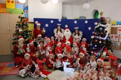 Przedszkolaki wraz ze świętym Mikołajem pozują do zdjęcia. W tle piękna dekoracja świąteczna. Przed nimi podświetlany renifer.