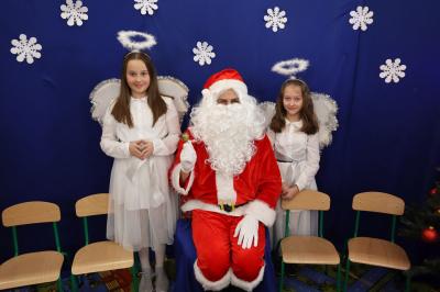 Mikołaj w asyście aniołków przemawia do dzieci. W tle piękna dekoracja świąteczna.