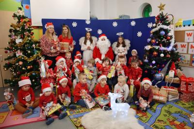 Przedszkolaki wraz ze świętym Mikołajem pozują do zdjęcia. W tle piękna dekoracja świąteczna. Przed nimi podświetlany renifer.