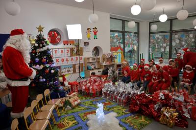 Przedszkolaki ubrane w czerwone stroje wpatrują się w Mikołaja. Przed nimi ułożone prezenty.