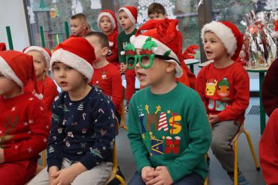 Święty Mikołaj podczas spotkania z przedszkolakami. Dzieci ubrane w mikołajkowe stroje śpiewają piosenki mikołajowi.