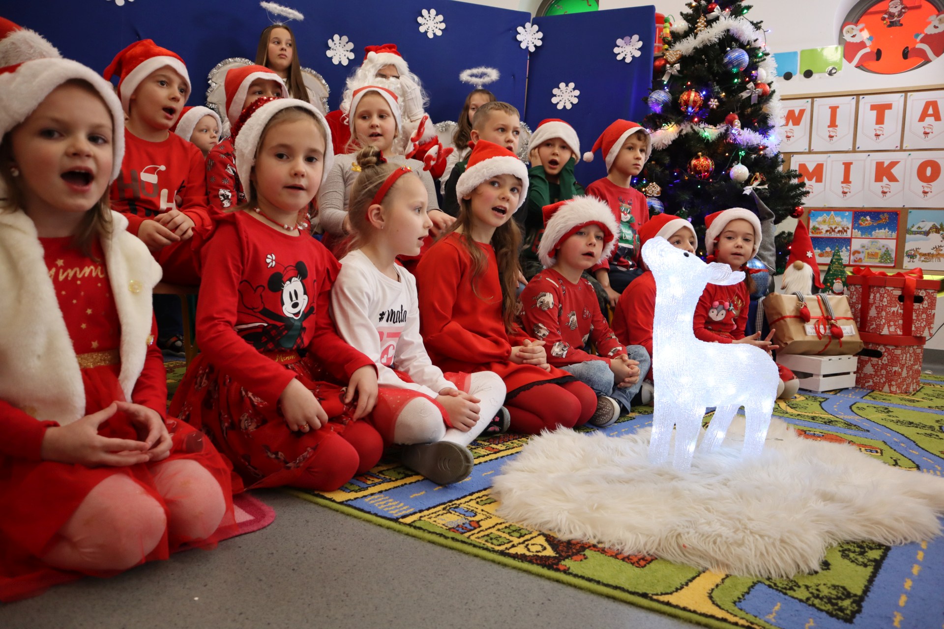Dzieci ubrane w czerwone stroje siedzą w czasie spotkania ze Świętym Mikołajem wokół podświetlanehgo reniferka.