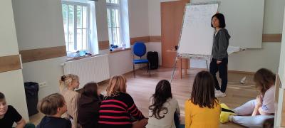 Uczestnicy obozu językowego,  chłopcy i dziewczynki siedząc na podłodze słuchają trenerki, która pokazuje im na planszy diagramy.
