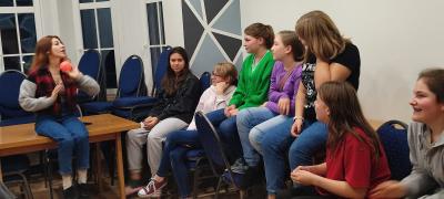 Dziewczynki siedząc na krzesłach bawią się z trenerką małą piłeczką, odpowiadają na pytania.