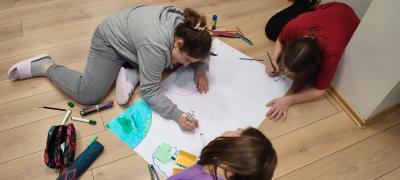 Dziewczynki leżąc na podłodze malują plakat z hasłem obozu EuroWeek.