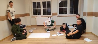 Chłopcy siedząc na podłodze malują plakat z hasłem obozu EuroWeek. Jeden z nich ma wyciętą z papieru maskę na twarzy.
