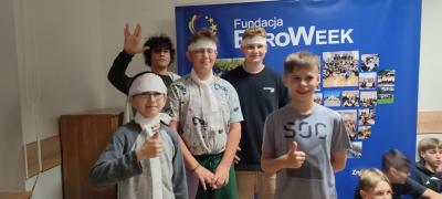 Wspólne zdjęcie uśmiechniętych uczestników projektu na tle plakatu EuroWeek. Chłopcy trzymają kciuki do góry.