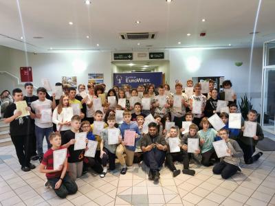 Grupowe zdjęcie wszystkich uczestników projektu na tle plakatu EuroWeek. Uczestnicy trzymają w rekach certyfikaty uczestnictwa w obozie językowym.